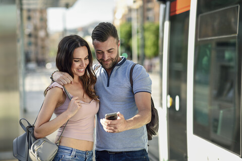 Ein Paar schaut auf sein Smartphone, während es am Bahnhof auf die Straßenbahn wartet, lizenzfreies Stockfoto