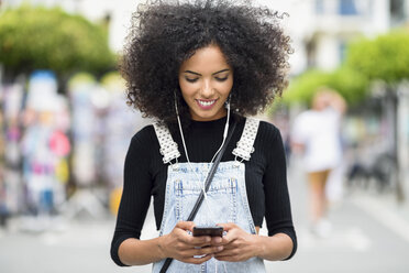 Smiling young woman with earphones looking at cell phone - JSMF00359