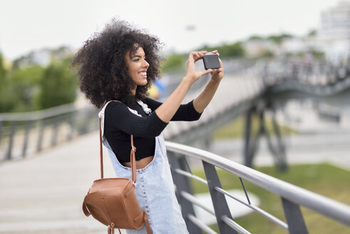 Lächelnde junge Frau mit braunem Lederrucksack, die auf einer Brücke steht und mit ihrem Smartphone Fotos macht - JSMF00370
