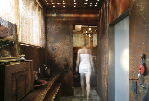 Rückansicht einer Frau, die ein Badezimmer mit Wandverkleidung aus Cortenstahl und Deckenlichteffekten betritt, lizenzfreies Stockfoto