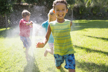 Drei Kinder jagen sich gegenseitig im Garten mit Wassersprinkler - CUF35281