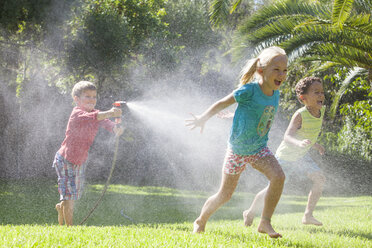 Three children in garden chasing each other with water sprinkler - CUF35282
