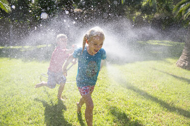Boy running after girl in garden with water sprinkler - CUF35284