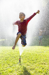 Boy jumping over water sprinkler in garden - CUF35286
