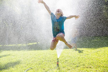 Girl jumping over water sprinkler in garden - CUF35287