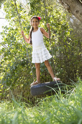 Girl standing swinging on tree tire swing in garden - CUF35342