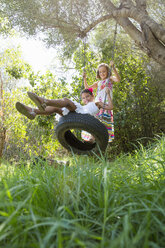 Two girls sitting and standing on tree swing in garden - CUF35343