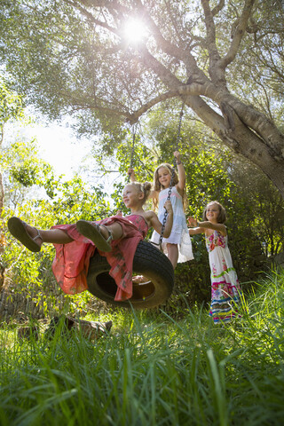 Niedriger Winkel Blick auf drei Mädchen spielen auf Baum Reifenschaukel im Garten, lizenzfreies Stockfoto