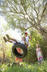 Low angle view of four girls playing on tree tire swing in garden - CUF35346