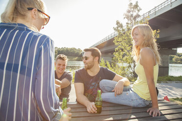 Vier junge Freunde trinken Bier auf einer Picknickbank am Flussufer - CUF35357