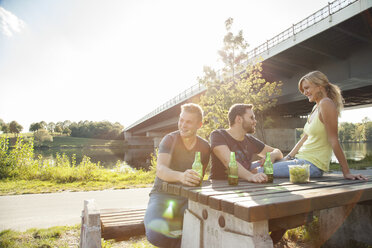 Drei junge Freunde trinken Bier auf einer Picknickbank am Flussufer - CUF35358