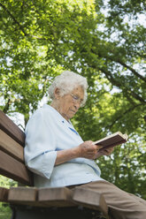 Ältere Frau sitzt auf einer Parkbank und liest in der Bibel - CUF35459