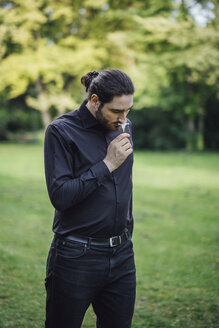 Young man, dressed in black, standing in park, smelling flower - JSCF00102