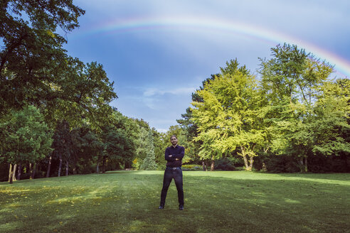 Young businessman standing in park under a rainbow, with arms crossed - JSCF00103