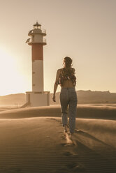 Spain, back view of young woman walking to lighthouse at sunset - ACPF00056