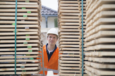 Porträt eines jungen männlichen Arbeiters zwischen Palettenstapeln auf einem Holzlagerplatz - CUF36213