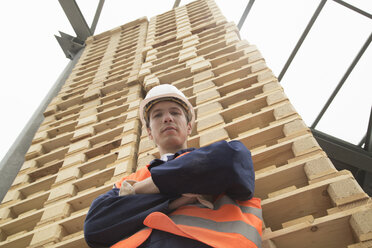 Low angle portrait of young man and tower of pallets in timber yard - CUF36214