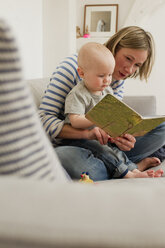 Reife Mutter liest ihrer kleinen Tochter auf dem Sofa im Wohnzimmer ein Bilderbuch vor - CUF36891