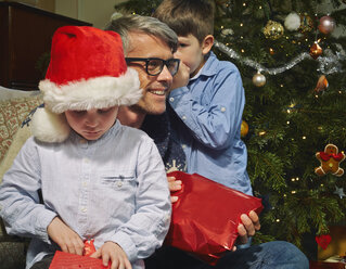 Boy whispering to father whilst brother opens christmas gift - CUF36932