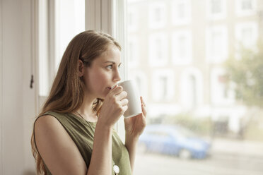 Young woman drinking coffee and gazing through sitting room window - CUF36933