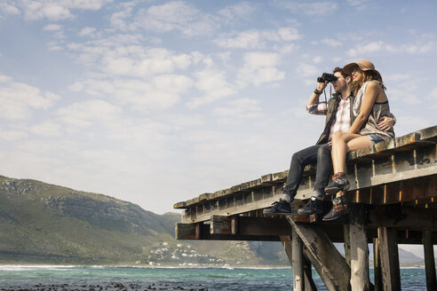 Junges Paar sitzt am Rand eines alten Piers und schaut durch ein Fernglas, Kapstadt, Westkap, Südafrika - CUF36949