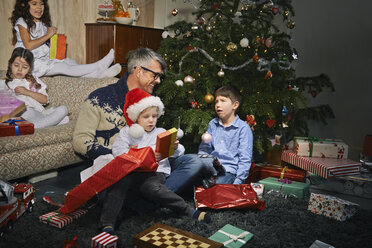 Vater im Wohnzimmer beim Öffnen der Weihnachtsgeschenke mit Kindern - CUF36951