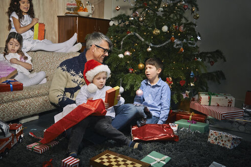 Vater im Wohnzimmer beim Öffnen der Weihnachtsgeschenke mit Kindern - CUF36951