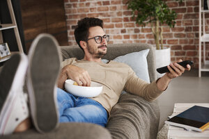 Content man lying on couch at home with bowl of potato chips watching TV - ABIF00655