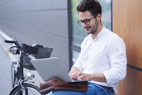 Businessman with bicycle using laptop outdoors stock photo