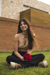 Porträt einer lächelnden jungen Frau mit roter Rose auf dem Rasen sitzend - ACPF00062