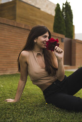 Lächelnde junge Frau sitzt auf dem Rasen und riecht an einer roten Rose - ACPF00063