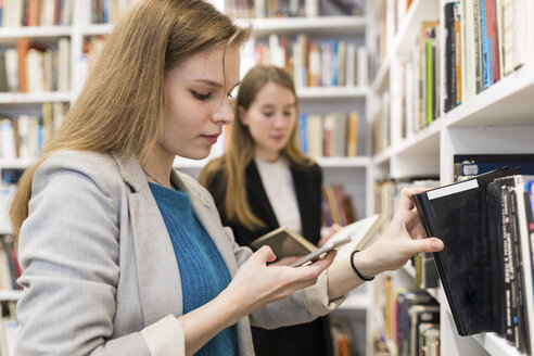 Porträt eines Teenagers, der auf sein Handy schaut, während er ein Buch aus dem Regal in einer öffentlichen Bibliothek nimmt - WPEF00490