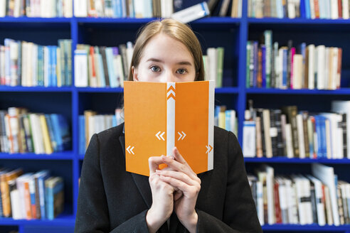 Teenage girl hiding behind book in a public library - WPEF00500