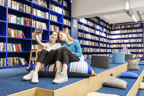 Zwei Teenager-Mädchen sitzen auf Sitzsäcken in einer öffentlichen Bibliothek und machen ein Selfie mit ihrem Smartphone, lizenzfreies Stockfoto