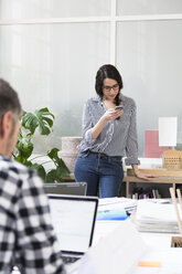 Porträt einer Frau mit einem Kollegen, der ein Mobiltelefon im Büro benutzt - FKF02961