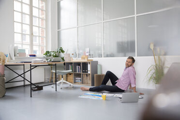 Casual man with plans and laptop sitting on the floor in a loft office - FKF02992