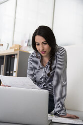 Lässige Frau mit Plänen und Laptop auf dem Boden sitzend im Büro - FKF02995