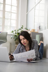 Casual woman with sheet of paper and laptop lying on the floor in office - FKF02998