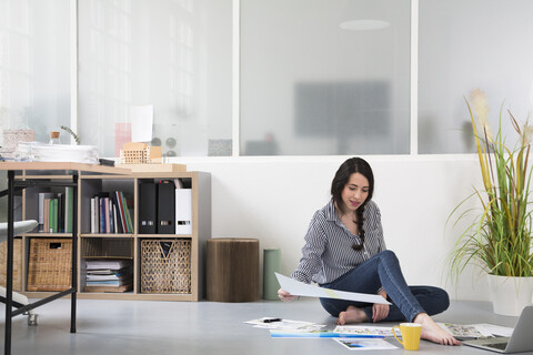 Lässige Frau mit Plänen und Laptop auf dem Boden in einem Loftbüro sitzend, lizenzfreies Stockfoto