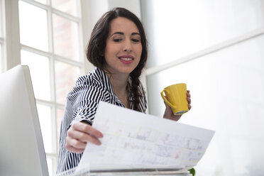 Smiling woman with cup of coffee looking at paper in office - FKF03024