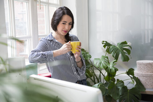 Lächelnde Frau mit einer Tasse Kaffee am Fenster im Büro - FKF03026