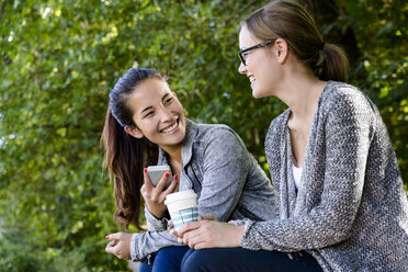 Zwei junge Frauen mit Kaffee zum Mitnehmen sitzen plaudernd im Park - CUF37592