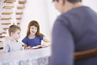 Großmutter beobachtet Enkelkinder beim Lesen eines Buches am Küchentisch - CUF37931