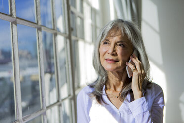 Senior woman looking out of window whilst using smartphone - ISF15181