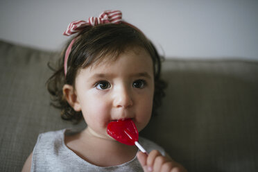 Cute baby girl eating a heart shaped lollipop at home - GEMF02100