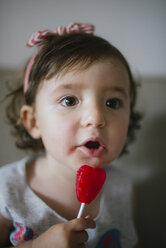 Cute baby girl eating a heart shaped lollipop at home - GEMF02102