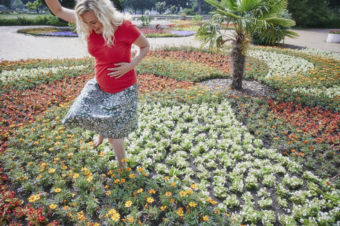 Happy pregnant woman walking through blooming flowers in park - RHF02054