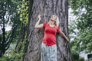 Smiling pregnant woman standing at a tree in park looking up - RHF02060