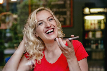 Portrait of laughing blond woman using smartphone in a cafe - RHF02075