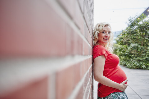 Porträt einer lächelnden schwangeren Frau, die sich im Freien an eine Backsteinmauer lehnt, lizenzfreies Stockfoto